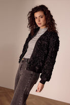 Woman wearing a black floral jacket over a gray sweater and jeans against a beige wall.