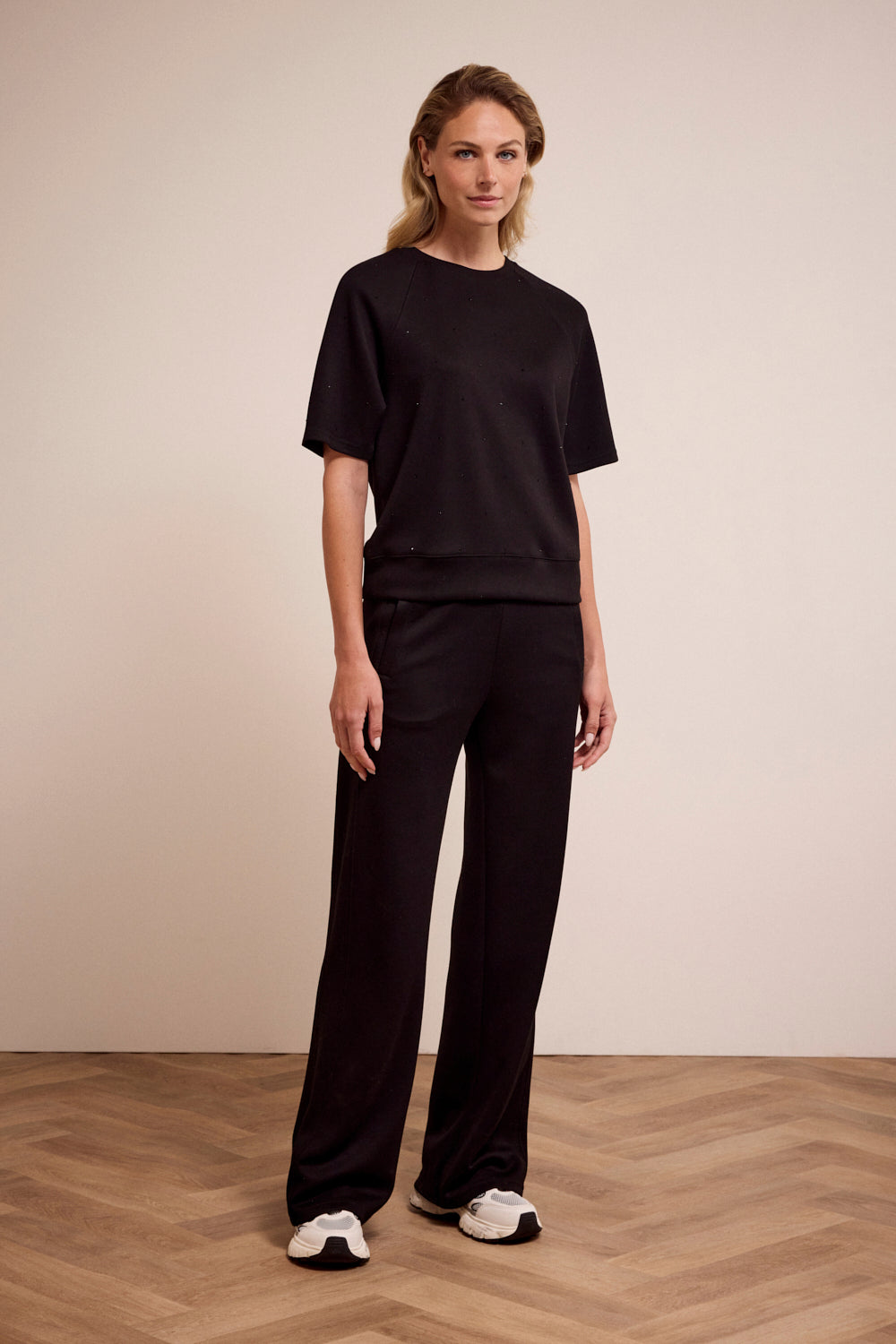 Woman wearing a black outfit standing on a wooden floor with a beige wall background