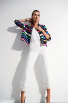 Woman wearing colourful blazer and white cropped pants styled with white heels, posing against a white backdrop.
