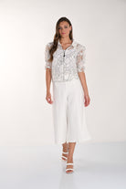Woman wearing white wide-leg culotte pants styled with a floral zip-front top and braided heeled sandals against a neutral studio background.