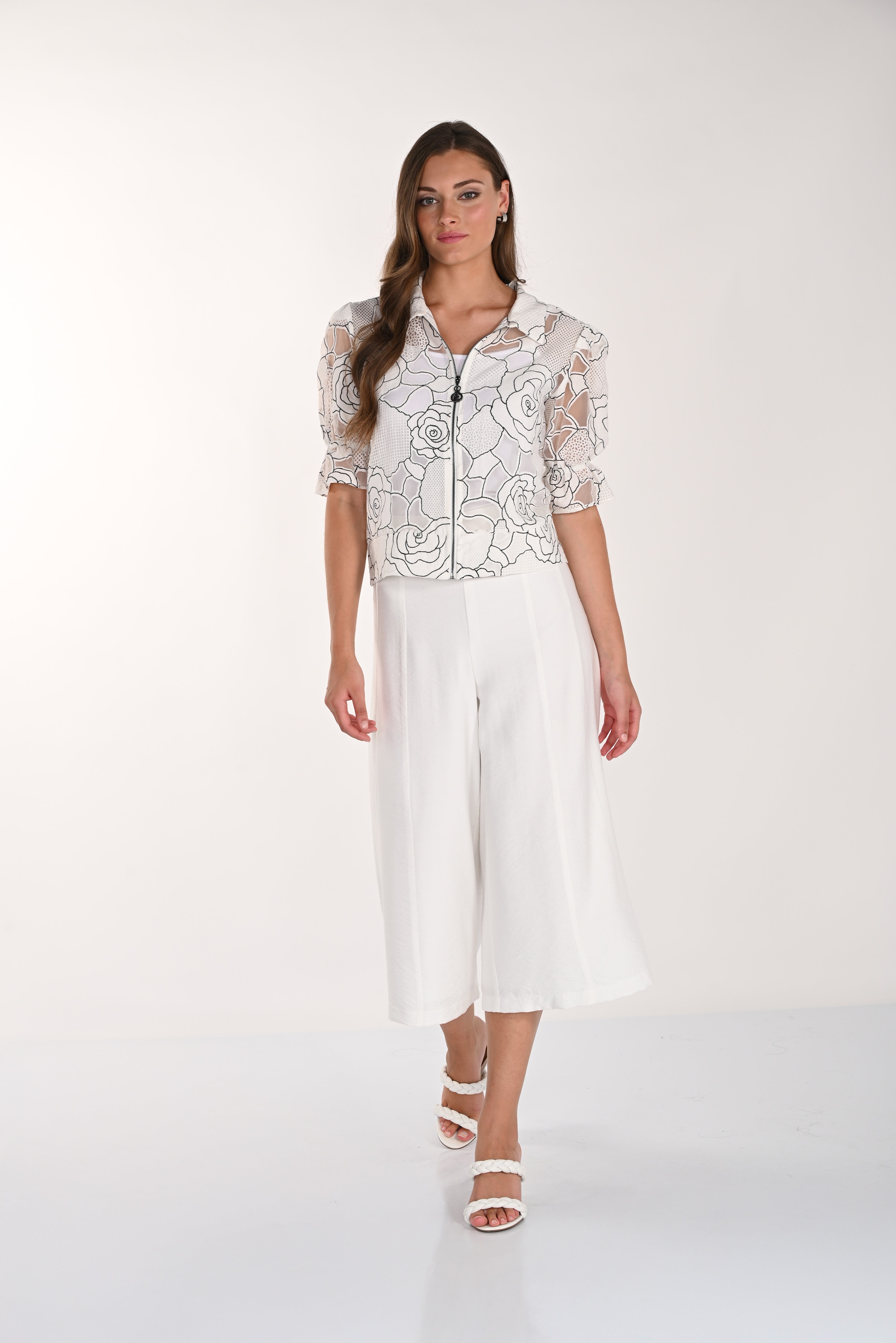 Woman wearing white wide-leg culotte pants styled with a floral zip-front top and braided heeled sandals against a neutral studio background.