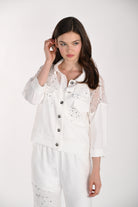 Model wearing a white button-front jacket with lace yoke detailing, sequin accents, patch pockets, and three-quarter sleeves, paired with matching trousers.
