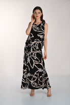 Front view of a women’s black maxi dress with an abstract beige floral print, featuring a sleeveless layered overlay design and flowing silhouette paired with heeled sandals.