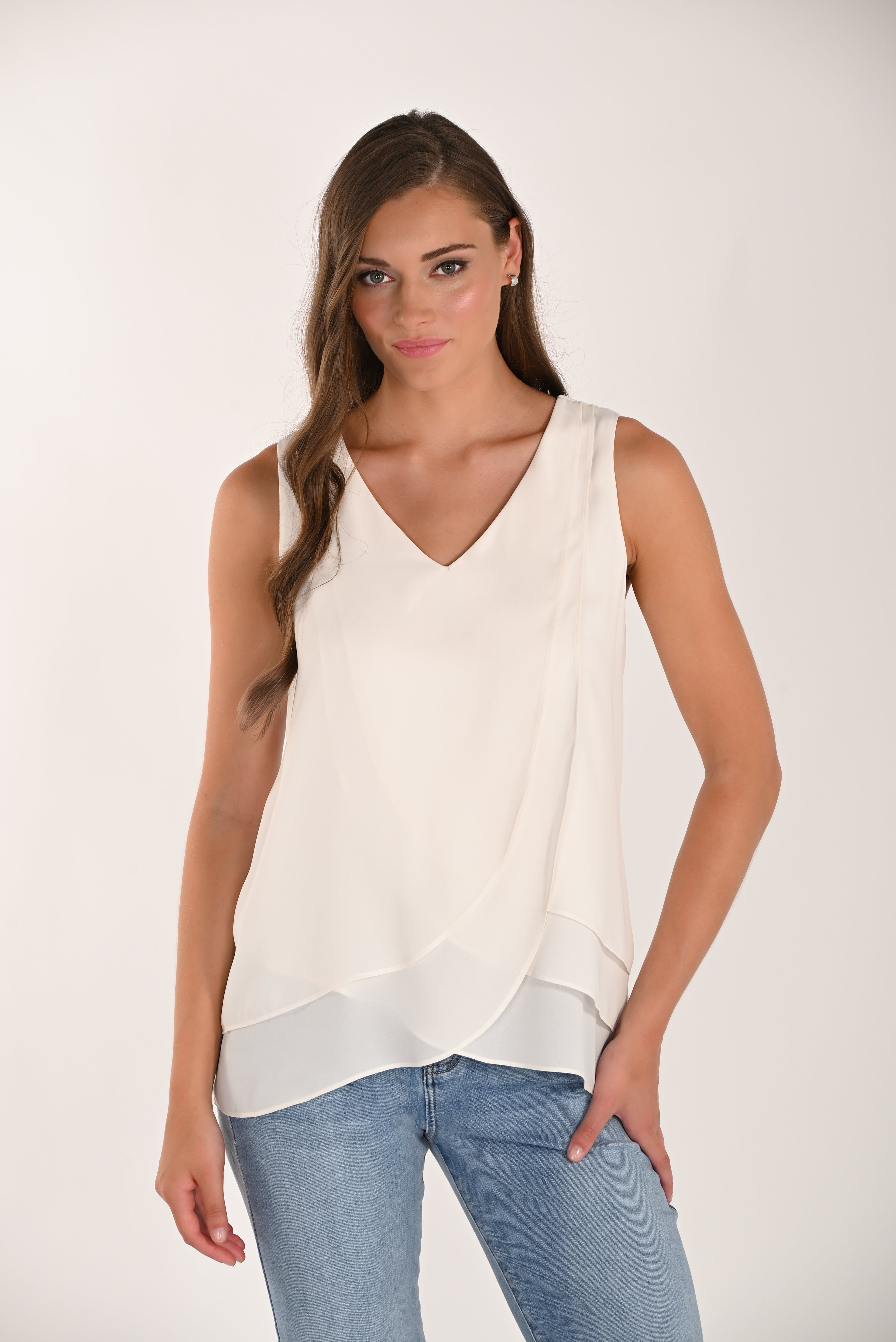 Woman wearing a sleeveless layered drape top with a V-neckline and flowing asymmetrical overlay, paired with light-wash jeans.