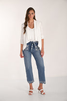 Model wearing medium-wash embroidered cropped wide-leg denim pants with a white layered blouse and white braided heeled sandals, featuring floral embroidery and scalloped hems for a feminine casual look.