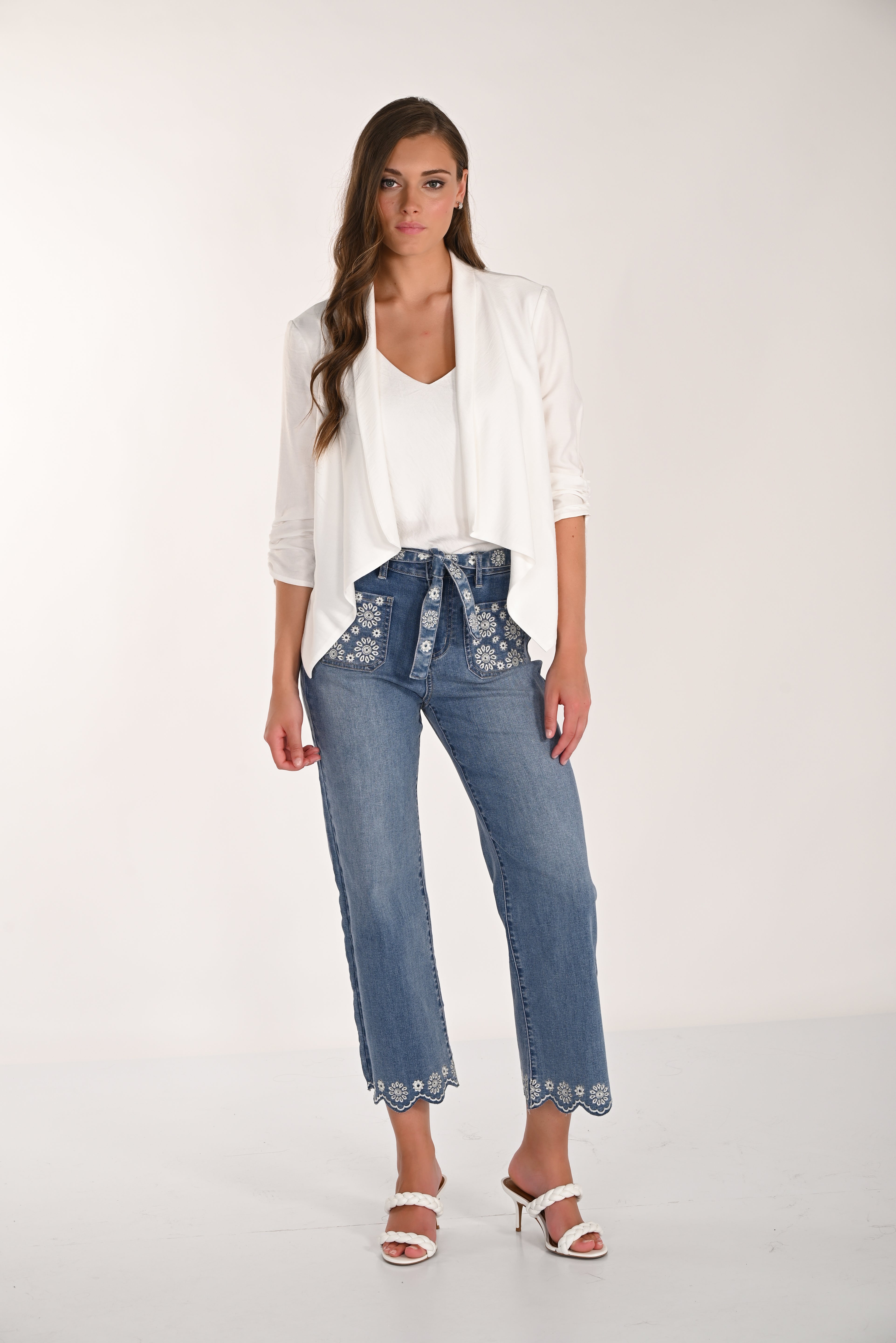 Model wearing medium-wash embroidered cropped wide-leg denim pants with a white layered blouse and white braided heeled sandals, featuring floral embroidery and scalloped hems for a feminine casual look.