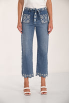 Woman wearing medium-wash embroidered cropped wide-leg denim pants with a tie waist, floral embroidery on the front pockets and waistband, and scalloped embroidered hems, styled with white braided heeled sandals.