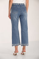 Back view of medium-wash cropped wide-leg denim pants featuring floral embroidery along the waistband and scalloped embroidered hems, paired with white braided heeled sandals.