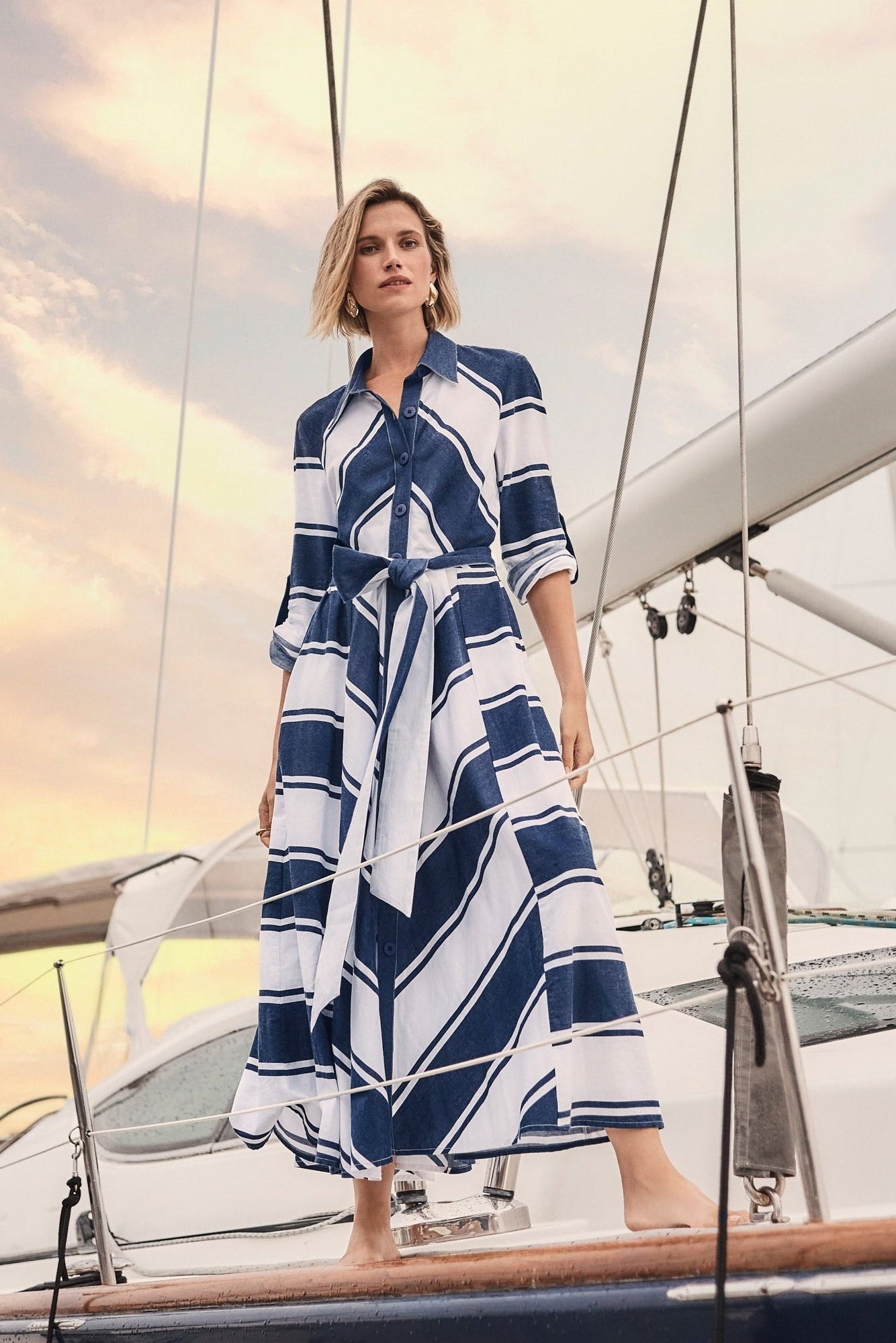 Model wearing Striped Cotton Fit and Flare Maxi Dress standing on a sailboat at sunset, showcasing bold navy and white stripes.