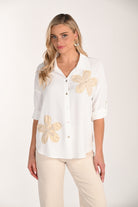 Woman wearing a white button-front roll-sleeve blouse with beige floral appliqué accents, paired with neutral trousers in a studio setting.