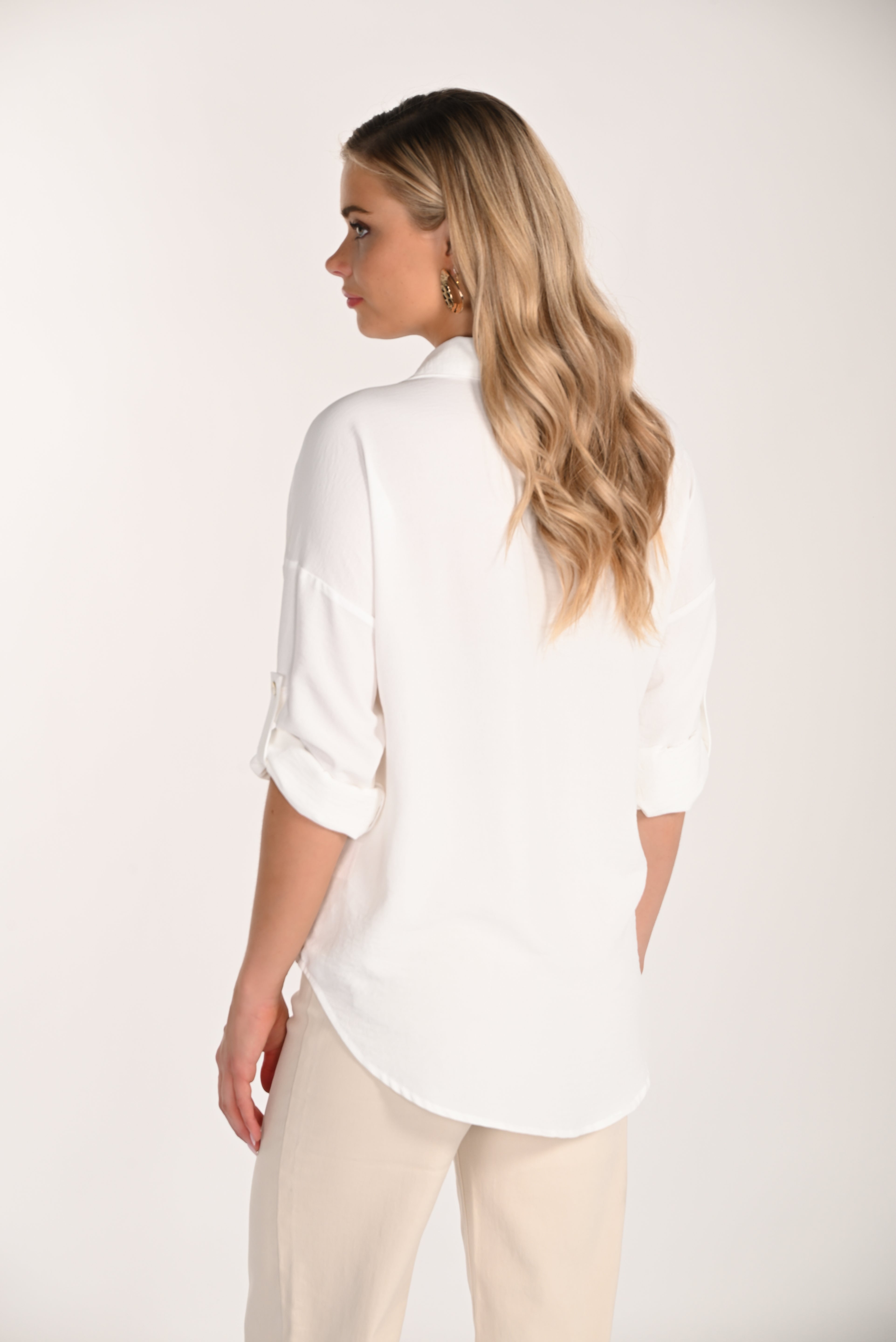 Woman wearing a white button-front roll-sleeve blouse with beige floral appliqué accents, paired with neutral trousers in a studio setting.