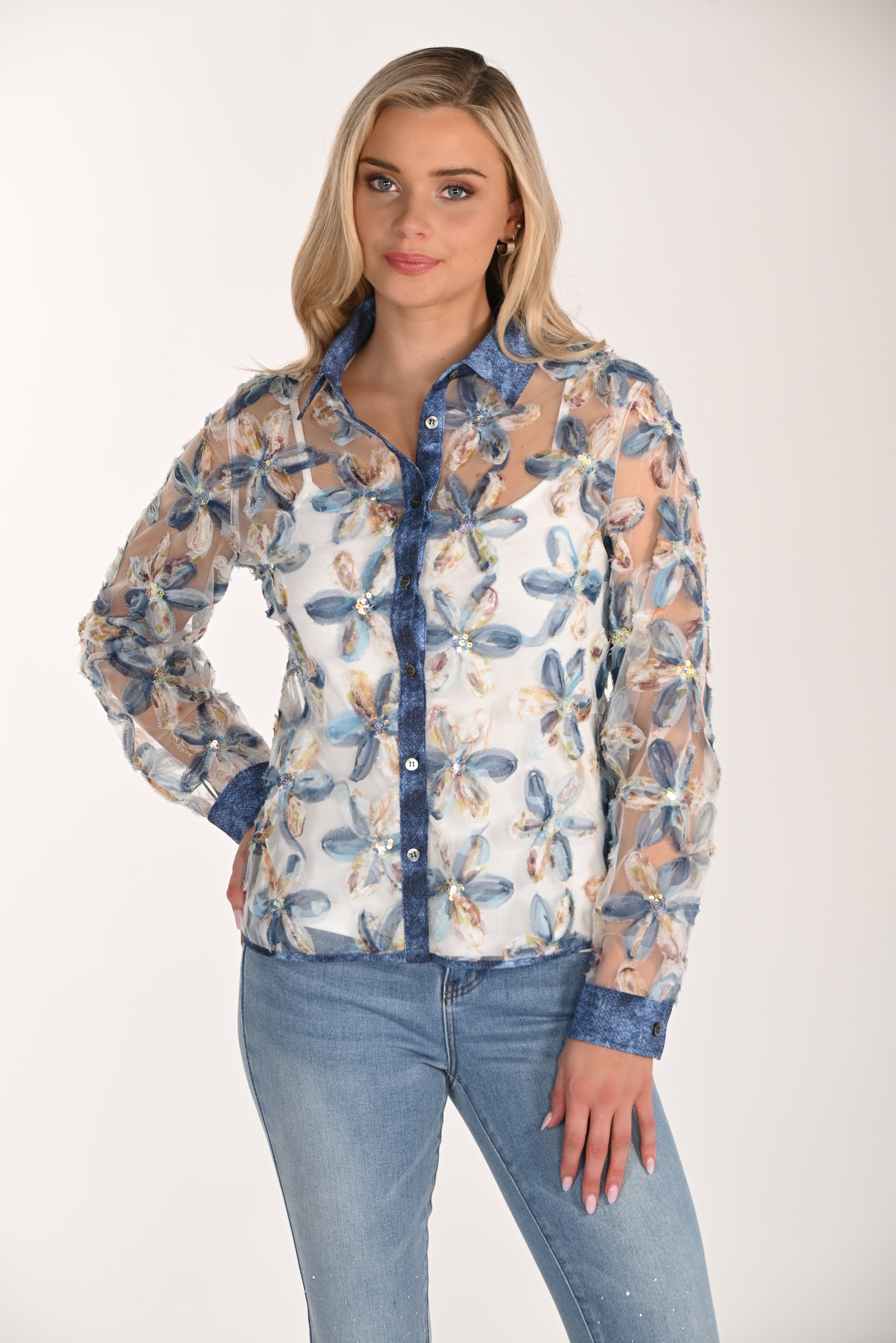 Front view of a women’s sheer floral mesh button-front blouse with textured blue and ivory embroidered flowers, long sleeves, and a collared neckline, styled with light wash jeans and a white camisole underneath.
