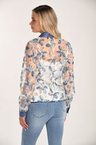 Back view of a women’s sheer floral mesh blouse with long sleeves, featuring textured blue and ivory embroidered flowers layered over a white camisole, paired with light wash jeans.
