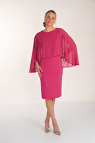 Front view of a bright pink knee-length sheath dress with a draped chiffon cape overlay and delicate shimmer accents, paired with silver heeled sandals.