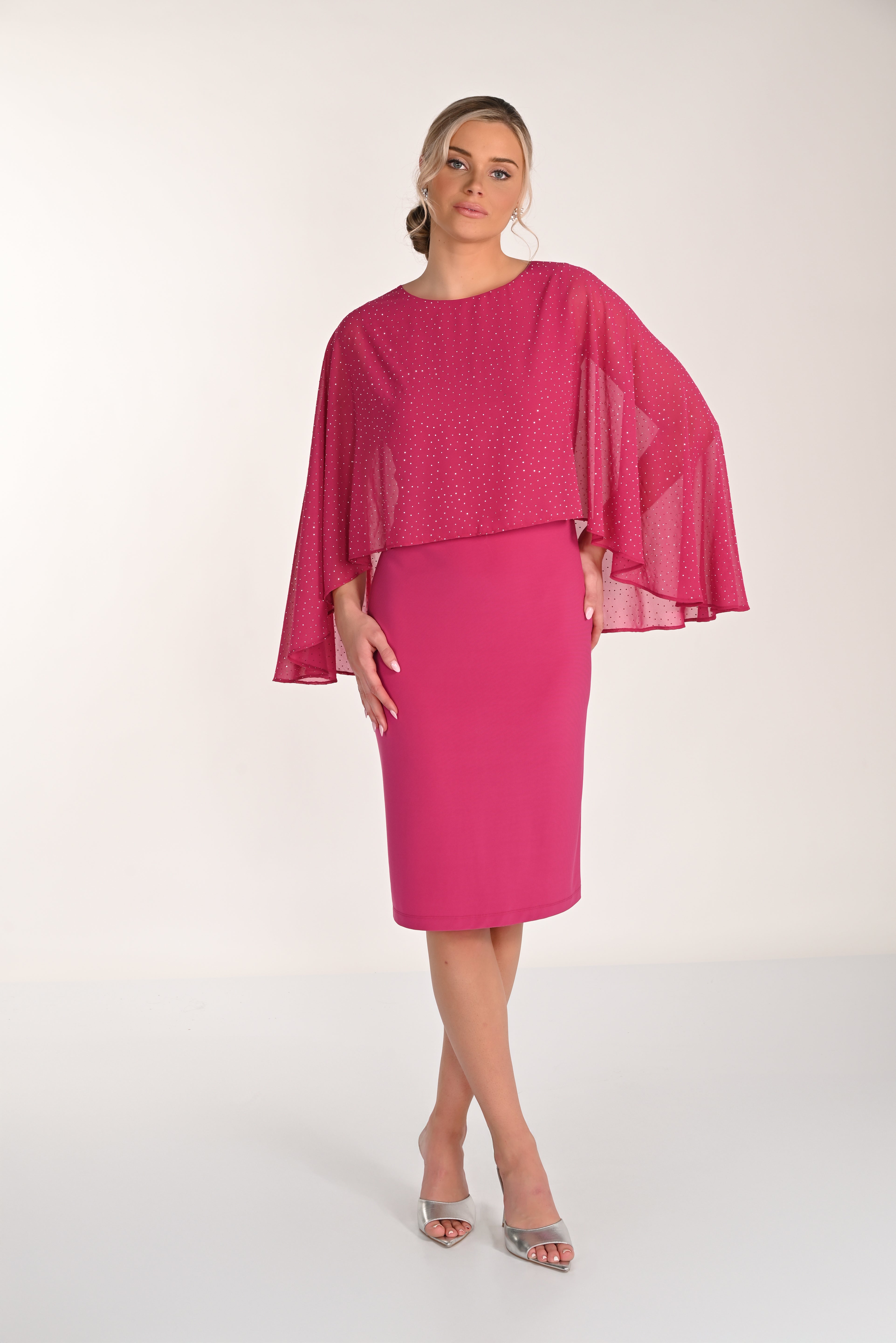 Front view of a bright pink knee-length sheath dress with a draped chiffon cape overlay and delicate shimmer accents, paired with silver heeled sandals.