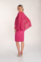 Back view of a bright pink knee-length sheath dress with a flowing chiffon cape overlay featuring subtle shimmer detailing, styled with silver heeled sandals.