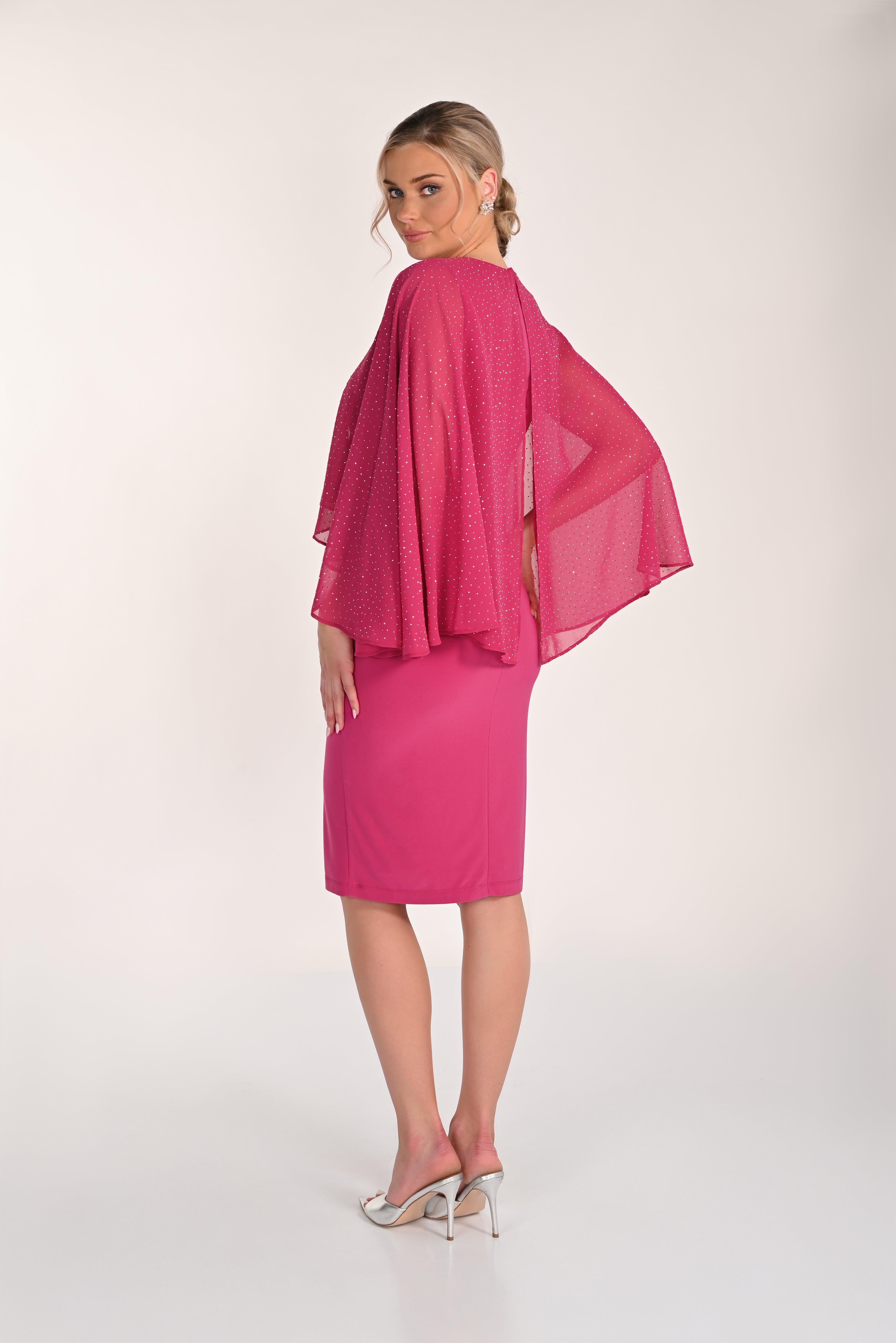 Back view of a bright pink knee-length sheath dress with a flowing chiffon cape overlay featuring subtle shimmer detailing, styled with silver heeled sandals.