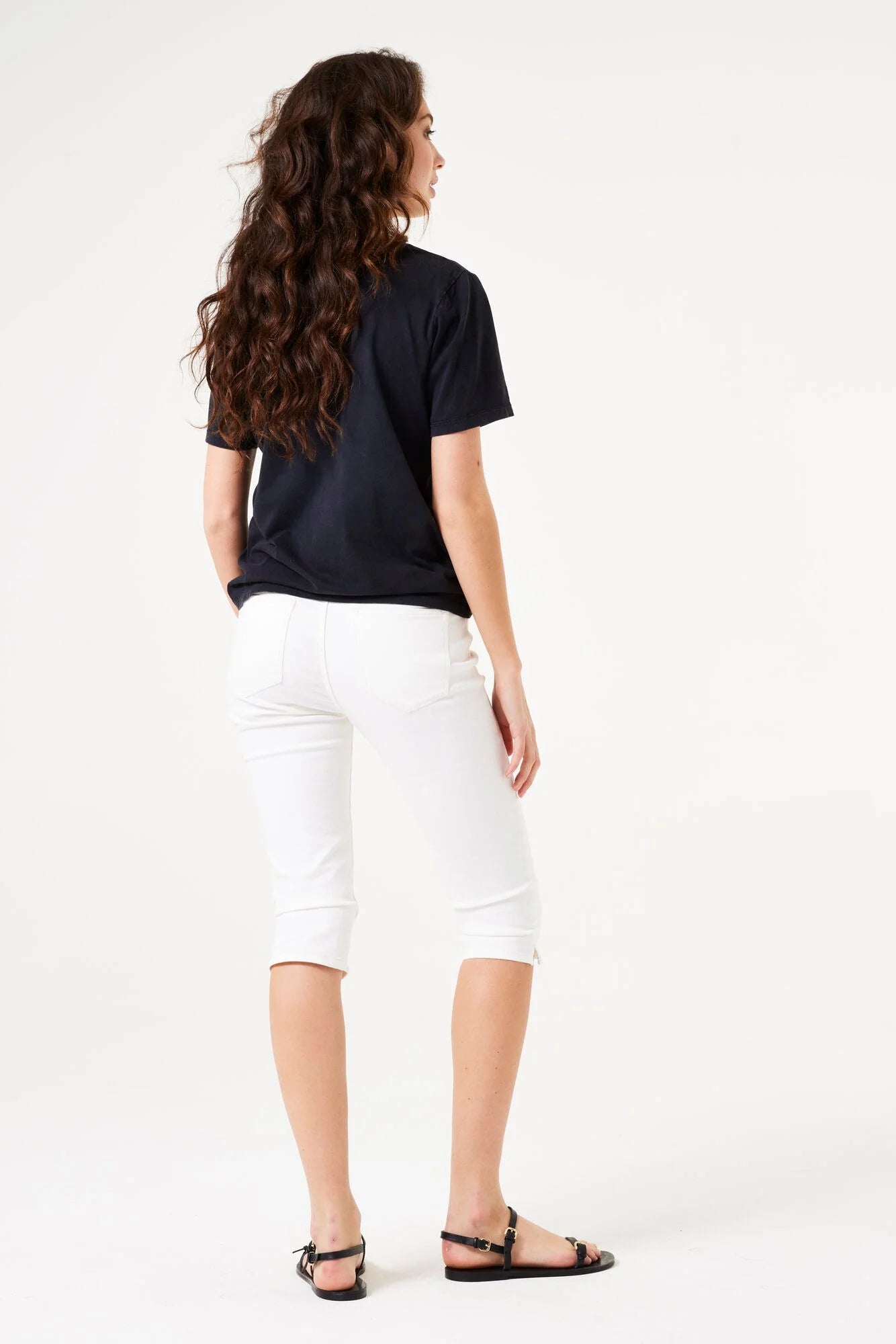 Back view of a woman wearing fitted capri pants with a cropped length, styled with a short-sleeve top and flat sandals.