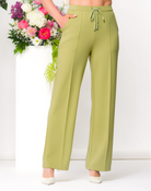 Woman with dark hair wearing a lime green wide-leg lounge pants with a drawstring waist, standing in a minimalist studio with floral arrangements behind her