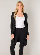 Women's black longline cardigan with an open-front design and long sleeves. Shown from the front, the cardigan is layered over a white tank top and black leggings, highlighting its soft drape and comfortable fit.
