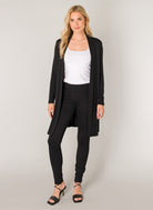 Women's black open-front longline cardigan styled over a white scoop-neck tank top and black leggings. The lightweight cardigan features long sleeves and a fluid drape, shown in a full-length front view with strappy heeled sandals.