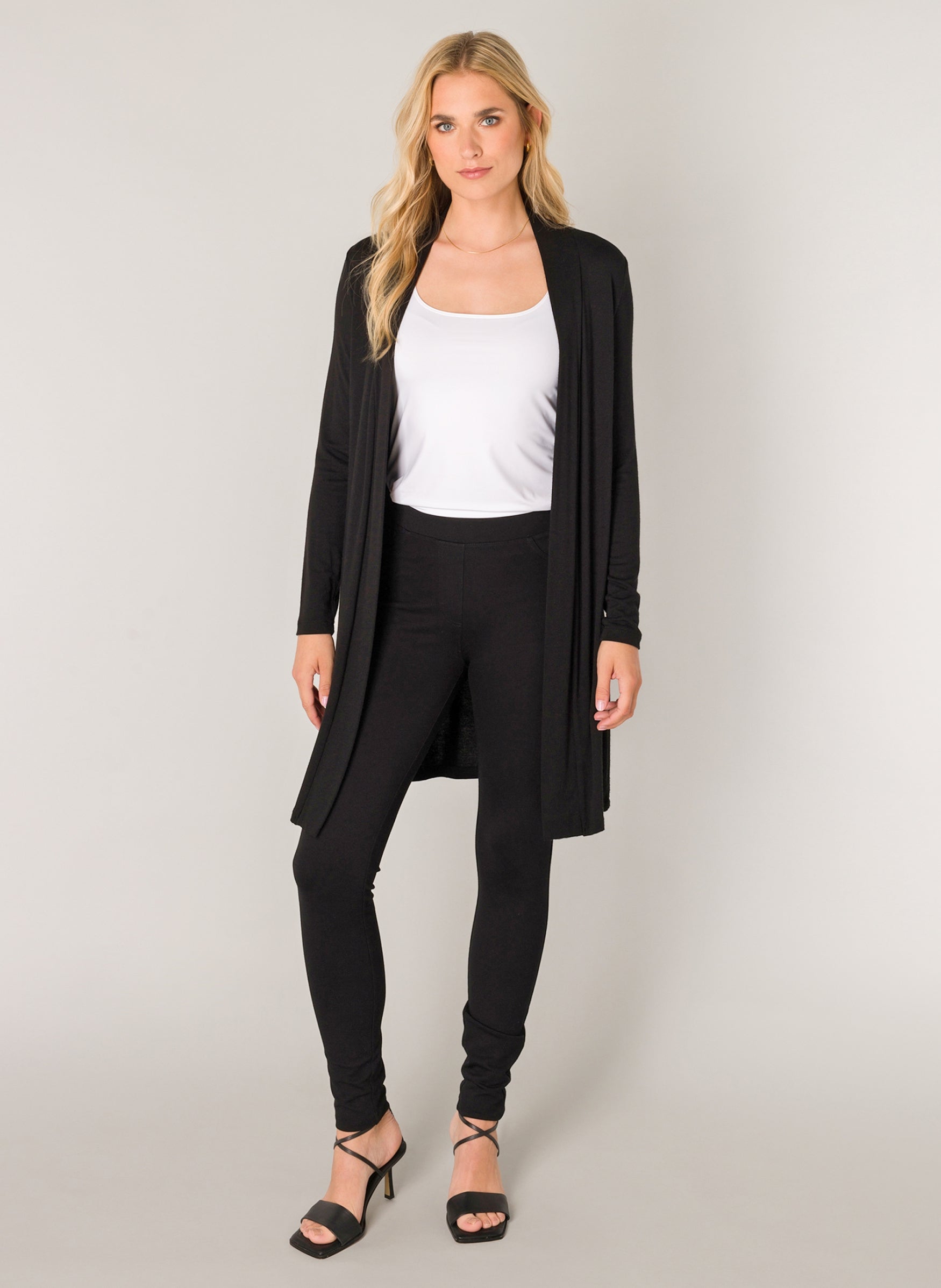 Women's black open-front longline cardigan styled over a white scoop-neck tank top and black leggings. The lightweight cardigan features long sleeves and a fluid drape, shown in a full-length front view with strappy heeled sandals.
