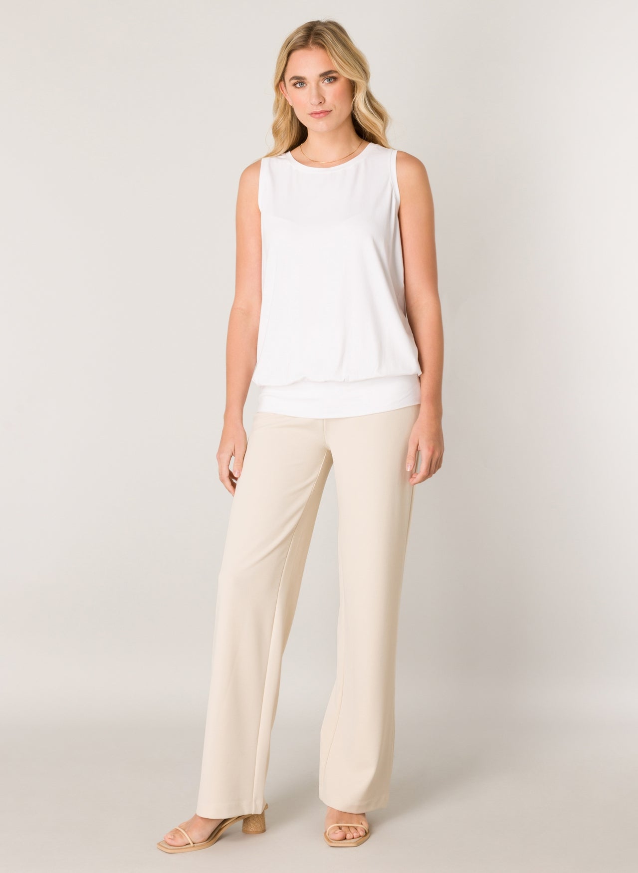 Women's white sleeveless blouse with a relaxed fit, round neckline, wide waistband hem, and soft draped silhouette, paired with cream wide-leg pants.