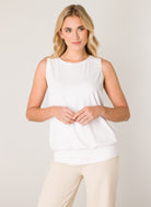Women's white sleeveless blouse with a relaxed fit, round neckline, wide waistband hem, and soft draped silhouette, paired with cream wide-leg pants.