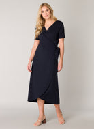 Women's dark blue wrap midi dress with a V-neckline, short sleeves, and self-tie waist detail. The soft stretch fabric drapes elegantly through the skirt, creating a flattering silhouette. Shown styled with strappy heeled sandals.