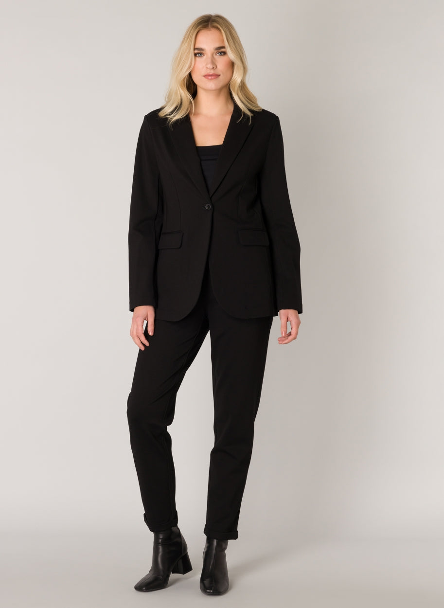 Full-length view of a model wearing a black tailored blazer with matching black trousers, featuring a notch lapel and single-button front closure.