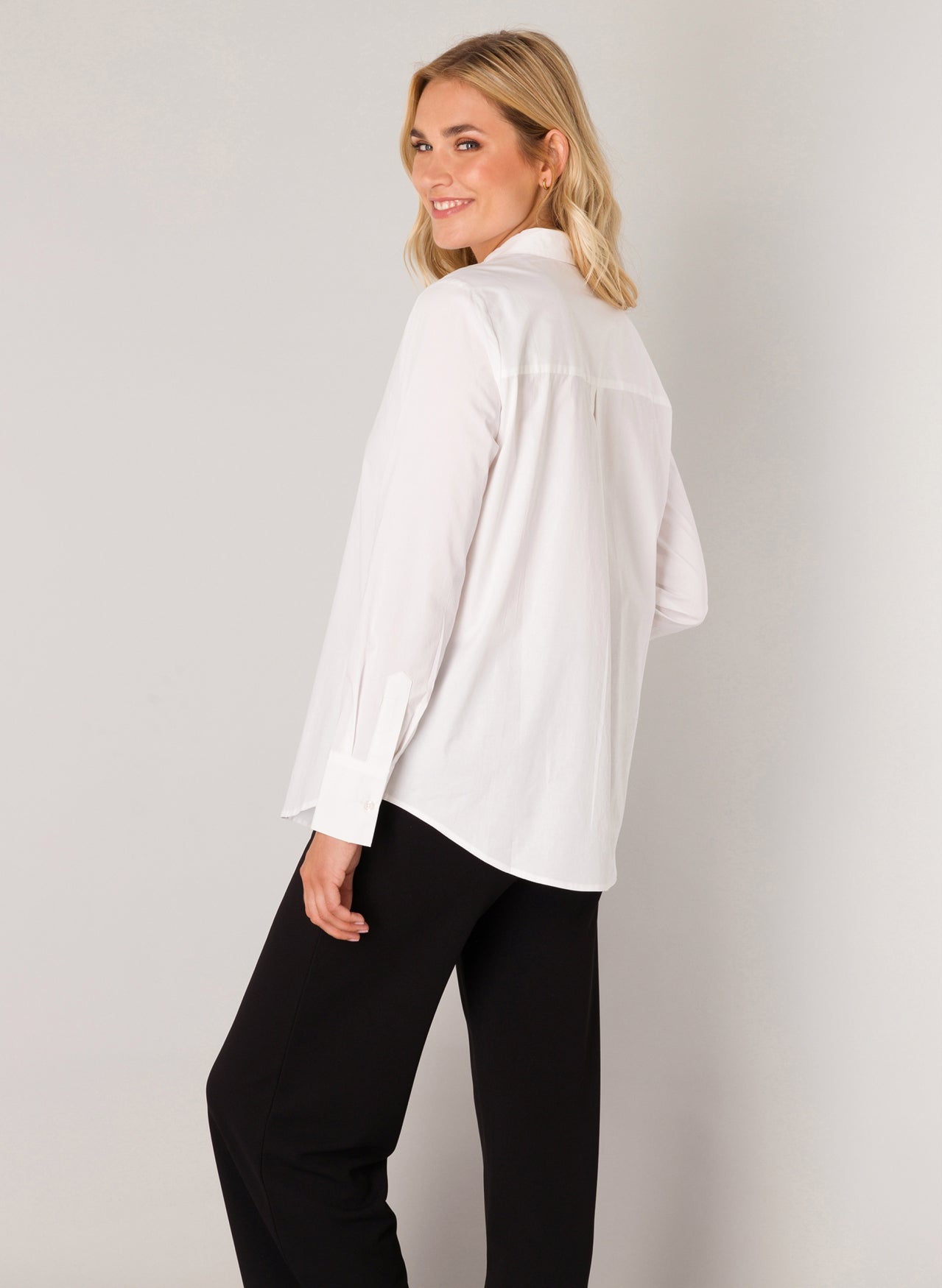 Back view of a woman wearing a white long-sleeve button-up shirt with a relaxed fit, paired with black pants, highlighting the clean back yoke and curved hemline.