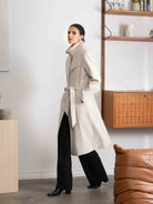 Woman wearing a long beige coat in a room with wooden shelves and a brown leather chair.