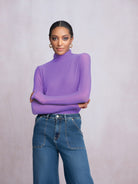 Woman wearing a purple turtleneck and blue jeans against a light gray background
