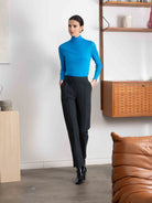 Woman in a blue top and black pants standing in a modern room with a brown leather sofa and wooden shelf