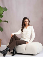 Woman sitting on a white ottoman wearing a white sweater and brown pants, with a plant in the background.