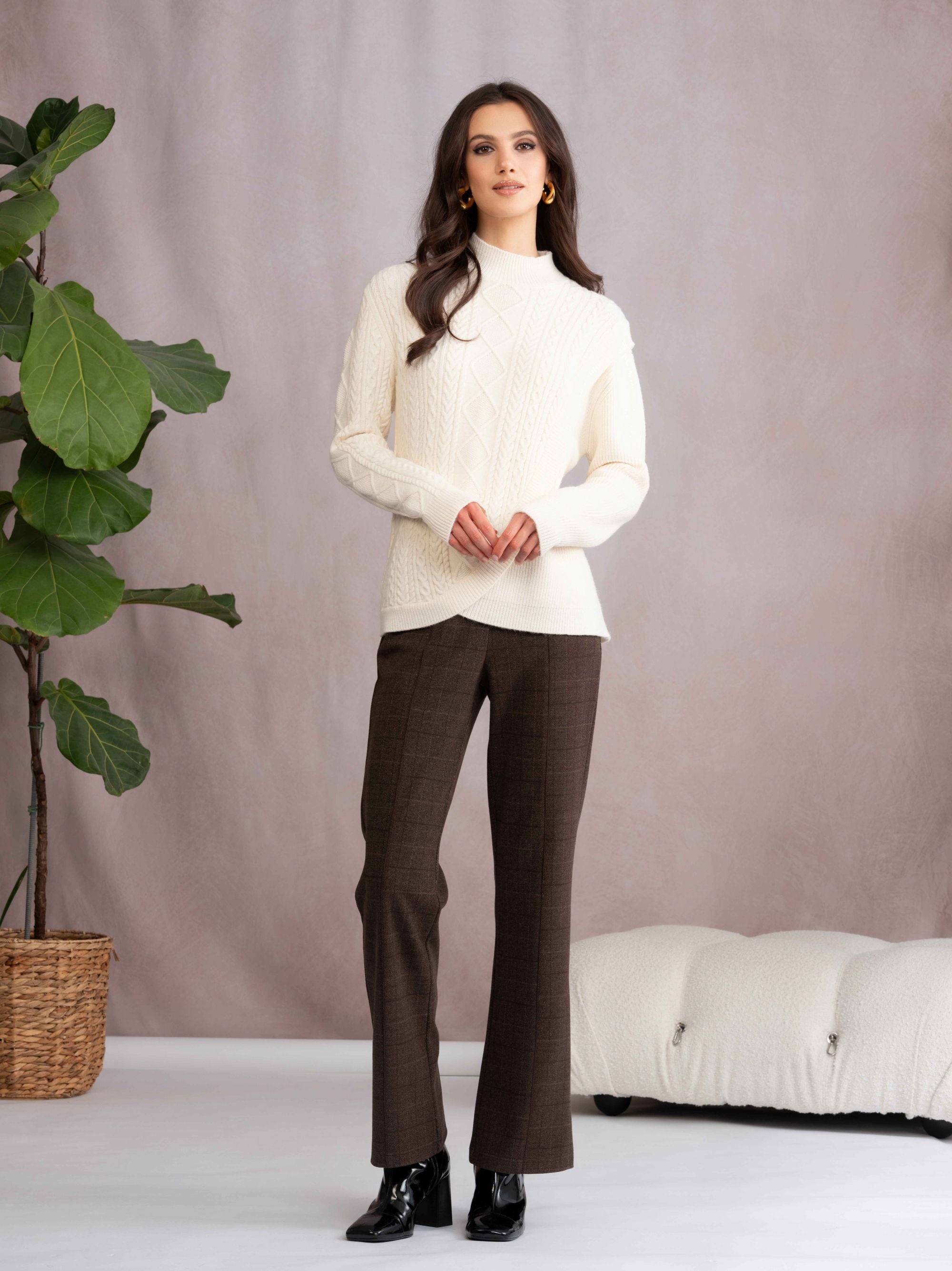 Woman wearing a white sweater and brown pants standing in a minimalistic room with a plant and sofa.