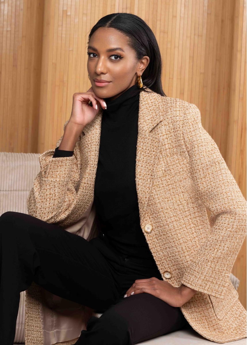 Woman wearing a beige blazer over a black outfit sitting on a couch with a wooden panel background