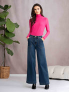 Woman wearing a pink turtleneck and blue jeans standing in a room with a plant and sofa.