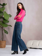 Woman wearing a pink sweater and blue jeans standing next to a plant indoors.