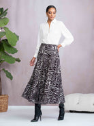 Woman wearing a white blouse and zebra print skirt standing indoors with a plant and white ottoman in the background.