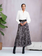 Woman wearing a white blouse and zebra print skirt with black boots, standing against a light purple wall.