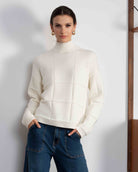 Woman wearing a white sweater and blue jeans standing against a white wall with a wooden shelf in the background.
