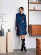 Woman in a blue patterned dress standing in a room with wooden shelves and a white wall.