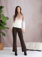 Woman wearing a gradient sweater and brown pants standing against a neutral background with a plant and white bench.
