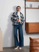 Woman in a plaid jacket and blue jeans standing in a room with wooden shelves and a brown leather chair.