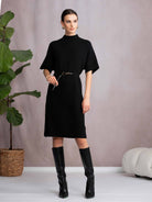 Woman wearing a black dress and boots standing next to a plant against a plain background