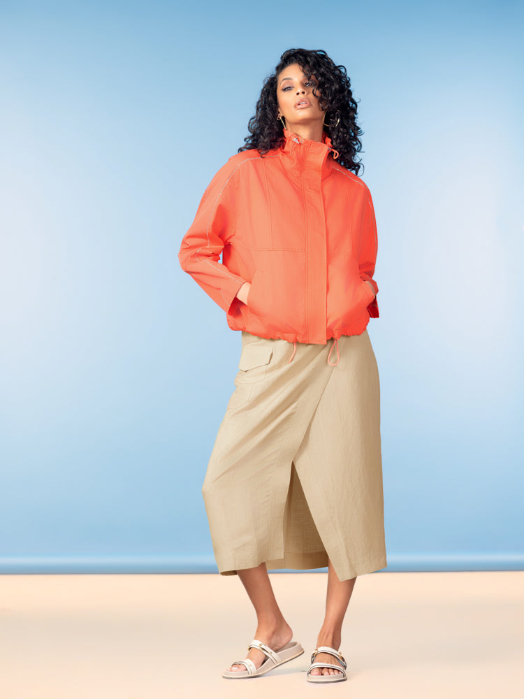Confident model wearing a lightweight coral zip-front jacket with a high collar and relaxed fit, paired with a sand-colored wrap midi skirt and neutral slide sandals, styled against a soft blue backdrop for a clean, modern look.