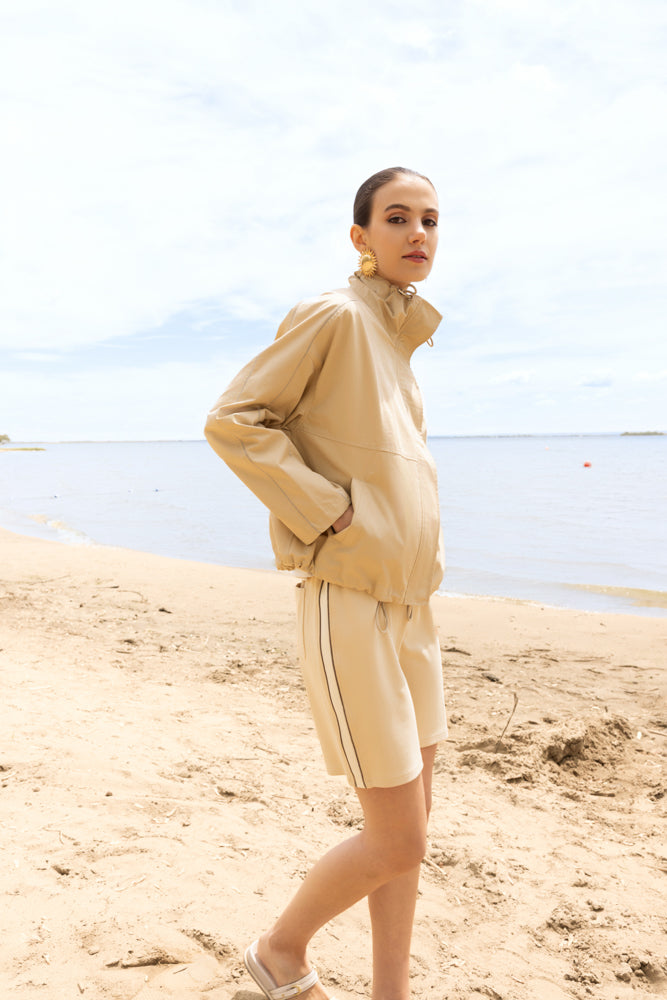 Woman wearing a zip-up light sand coloured jacket and matching shorts, standing in profile on a sandy beach with hands in pockets, styled in a relaxed, modern coastal outfit.