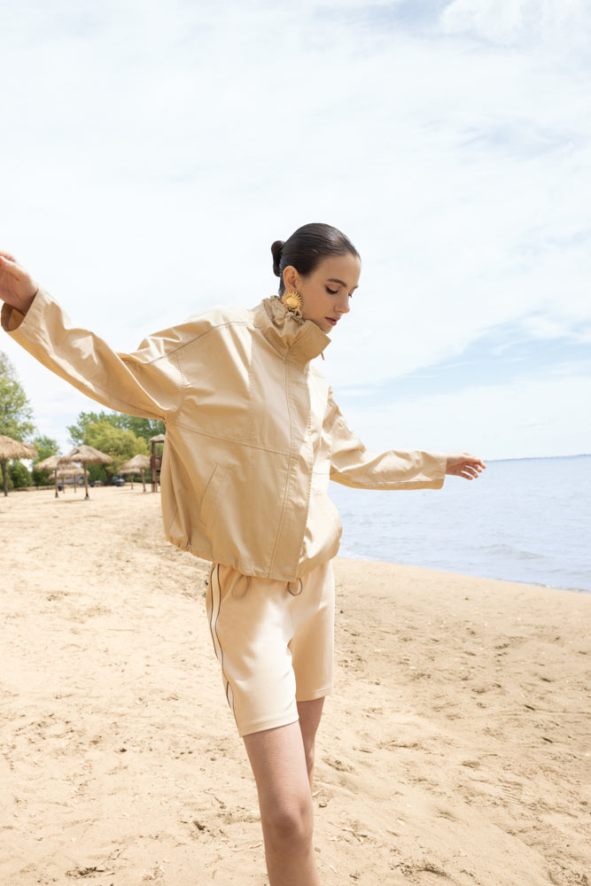 Model balancing gracefully along a sandy shoreline, wearing a sand-toned lightweight jacket with a high collar and matching tailored shorts. The monochrome look is styled with statement gold earrings, blending effortless elegance with coastal ease against a calm beach backdrop