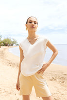 Woman in a white sleeveless top and beige shorts standing on a sandy beach with water and sky in the background.